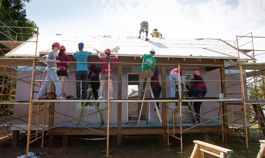 UA students wear hard hats and stand on ladders or the roof as they work on a house surrounded by scaffolding.