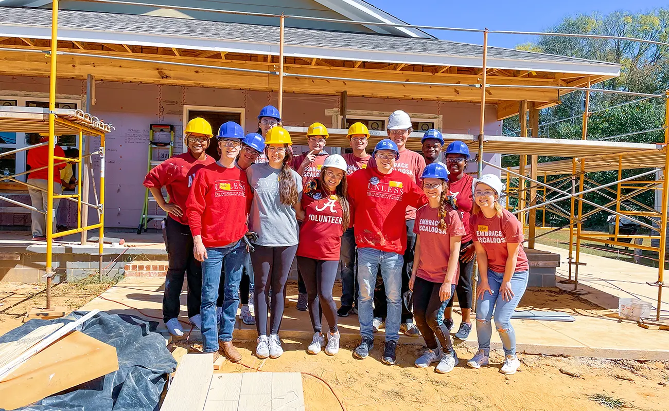 UA students wear hard hats and volunteer shirts, posing for a photo in front of a house they are building.