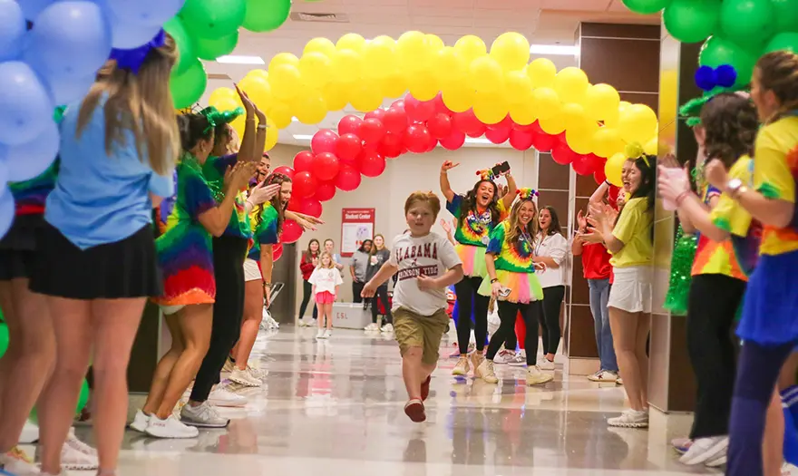 A child runs through various colors of balloon arches as UA Miracle teams of students cheer.