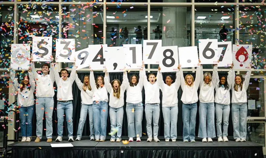 Students on a stage under falling confetti while holding signs displaying a fundraising total of $3,941,786.67.